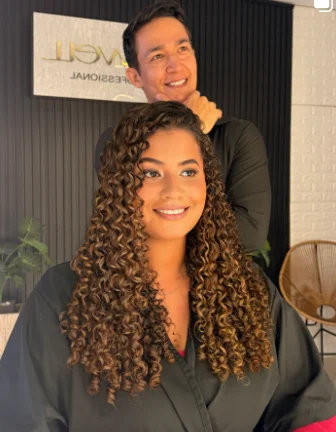 Cliente de cachos iluminados ao lado de Daniel Campos no salão
